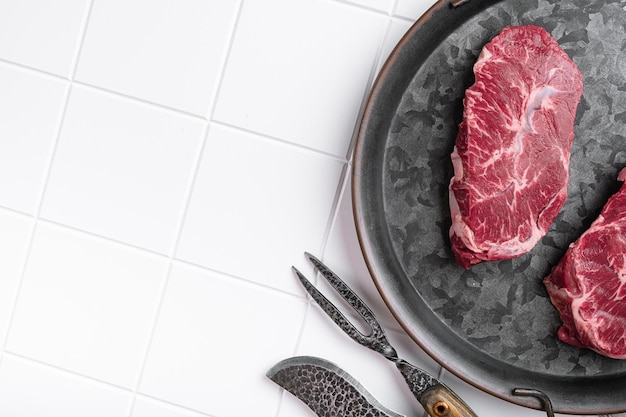 Marbled beef steak top blade meat steak on white ceramic\
squared tile table background top view flat lay with copy space for\
text