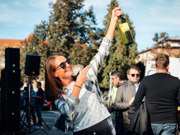 사진 마리보르, 슬로베니아 - 2019년 10월 11일: 선글라스를 끼고 웃고 있는 어린 소녀가 와인 병을 들고 있습니다. 여성 캐주얼 스타일입니다. 재미 여성 사람입니다. 행복한 휴일 축하. 야외 라이프 스타일 초상화입니다.