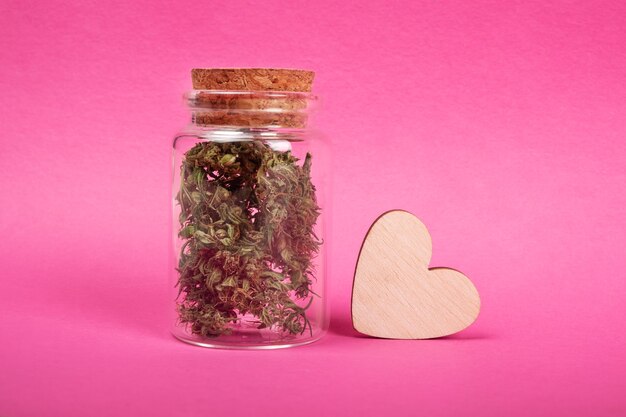 Marijuana love symbol jar with dry cannabis buds and a wooden heart on pink background