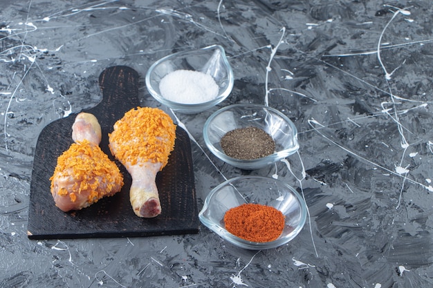 Marinated drumsticks on a cutting board next to spice bowls , on the marble background. 