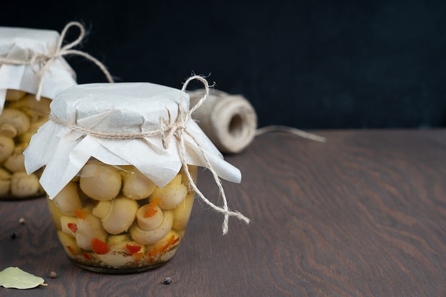 Marinated or fermented champignon mushrooms in glass can or jar on dark wooden table