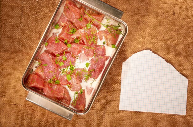 Foto carne marinata nel latte e verdure in un piatto fondo d'acciaio su un panno fatto in casa con fogli di carta puliti e una penna a sfera