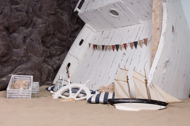 Морской или морской тематический натюрморт, изображающий затонувший корабль с деревянной лодкой, перевернутой в песок, с корабельным колесом, фонарем, опрокинутой игрушечной яхтой и разбросанными вокруг деревянных ящиками.