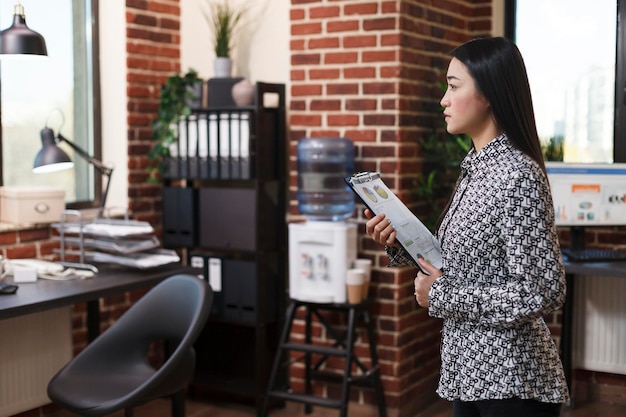 Marketing company office employee having project development chart in hand while looking towards desk. Young businesswoman holding clipboard in hands while standing in workspace.