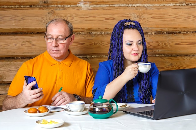 A married couple a man in glasses and a woman with blue afro braids at the table a woman drinks tea and works at a computer listens to a man. Horizontal photo