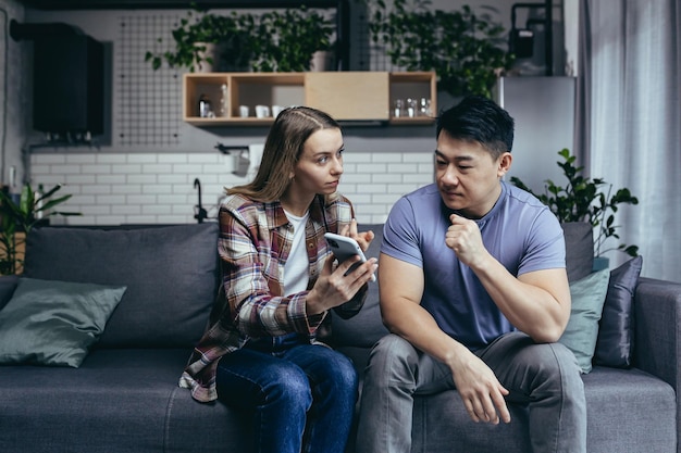 Married couple man and woman sitting on the couch at home and arguing over the phone jealousy