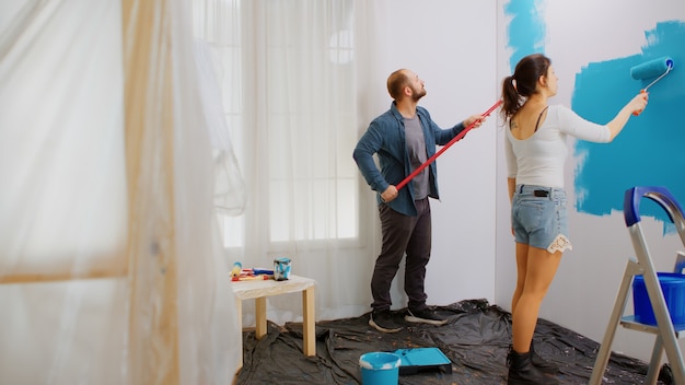 Married couple painting apartment wall with roller during home renovation using blue paint. Decorating, color, repair, decoration.