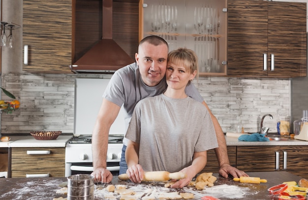 A married couple during the preparation of cookies stands near the table in the kitchen on which a lot of flour is scattered.