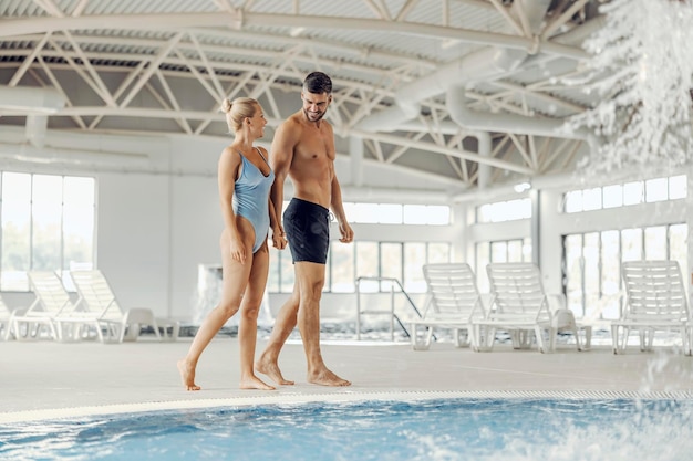 A married couple in swimwear is holding hands and walking next to a pool with thermal water