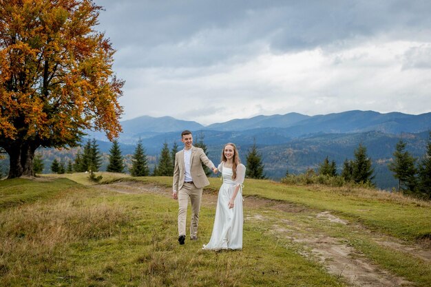 A married couple walks among the mountains the bride leads the groom behind her the girl holds the boy's hand smiles the woman in a white wedding dress the groom and a beige suit