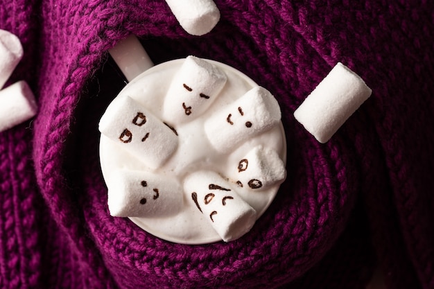 Marshmallows with painted faces in a mug with cappuccino wrapped in a knitted scarf