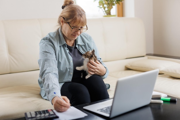 Marure woman in blue shirt sitting with a cat on her lap at the wooden table at home with laptop and notebook working or shopping online