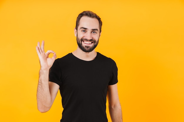 masculine man wearing basic black t-shirt smiling and gesturing ok sign isolated over yellow wall