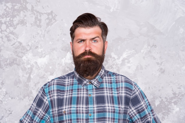 Masculine traits. Brutal bearded man concrete background. Serious man with beard and moustache. Confident hipster. Hairdresser in checkered shirt. Hipster style. Guy hipster outfit. Mature and trendy.