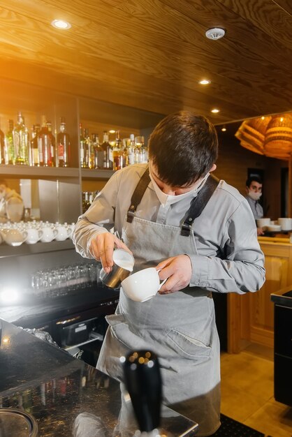 A masked barista prepares delicious coffee at the bar in a cafe. The work of restaurants and cafes during the pandemic.