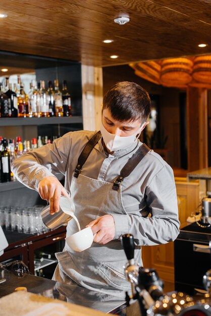 A masked barista prepares delicious coffee at the bar in a cafe. The work of restaurants and cafes during the pandemic.