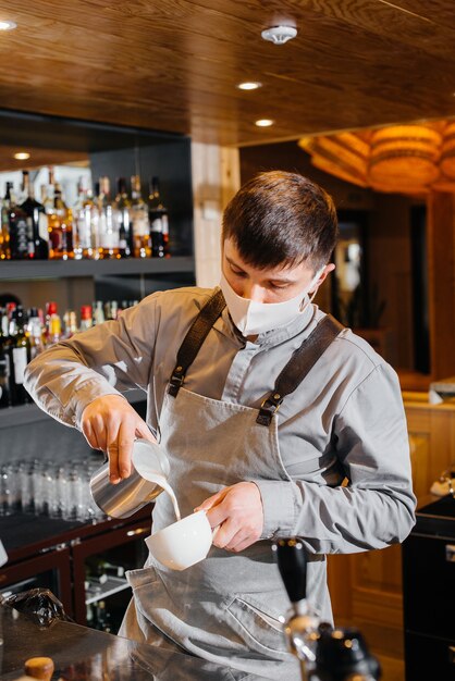 A masked barista prepares delicious coffee at the bar in a cafe