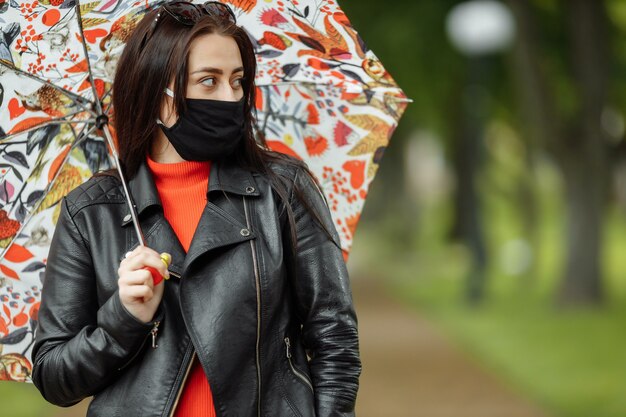 A masked girl is walking along the street. A girl in a protective mask walks in the park with an umbrella in the rain. Coronavirus infection COVID-19.