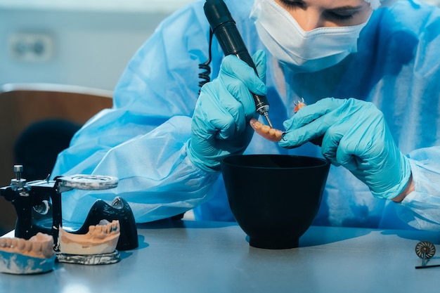 A masked and gloved dental technician works on a prosthetic tooth in his lab.