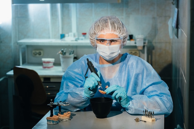 A masked and gloved dental technician works on a prosthetic tooth in his lab