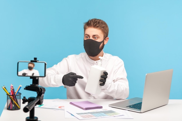 Masked and gloved man pointing bottle with antibacterial gel, showing antiseptic to webcam online broadcast, recommendations for hygiene, disinfection and safe office work during coronavirus pandemic