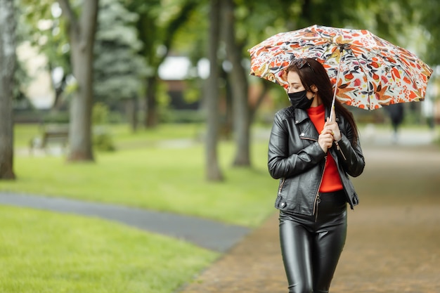 Женщина в маске идет по улице. Женщина в защитной маске гуляет в парке с зонтиком под дождем. Коронавирусная инфекция COVID-19