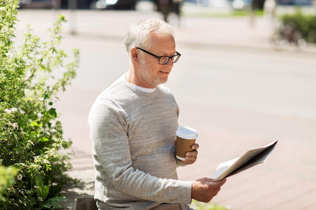 mass media, news and people concept - senior man reading newspaper and drinking coffee in city