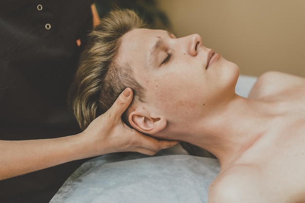 The masseur does a facial massage to a young guy. A man is massaged face on a massage table