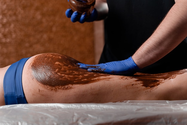 Il massaggiatore spalma di cioccolato le gambe e le cosce della ragazza nel centro termale. impacco al cioccolato con procedura di bellezza.