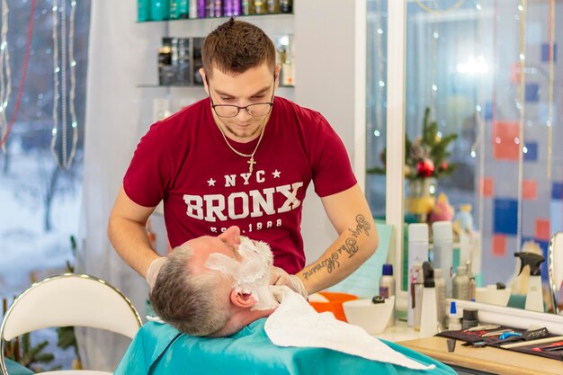 A master applies shaving cream to the beard of an adult man in a hairdressing salon with a shaving