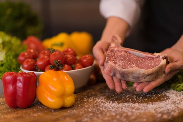 Master Chef holding juicy slice of raw steak with vegetables around on a wooden table