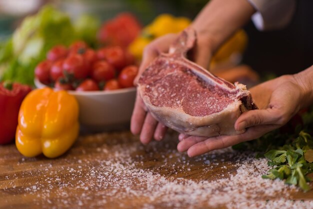 Master Chef holding juicy slice of raw steak with vegetables around on a wooden table