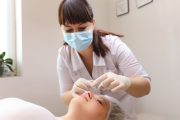 Мастер-косметолог наклоняется к клиентке и делает уколы в губы гиалуроновой кислотой для их увеличения.