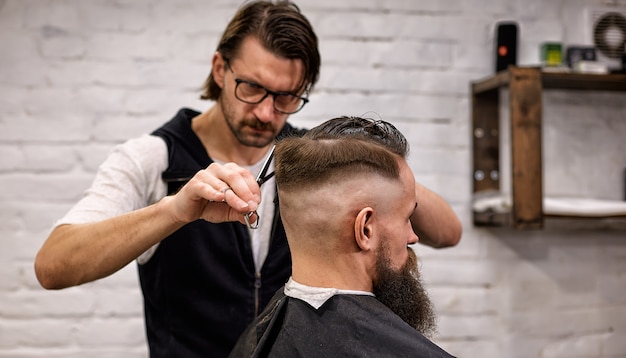 Мастер стрижет волосы и бороду мужчины в парикмахерской, парикмахер делает прическу для молодого человека