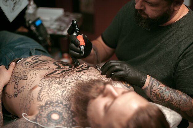 Master does tattoo on the skin of his client in tattoo parlour