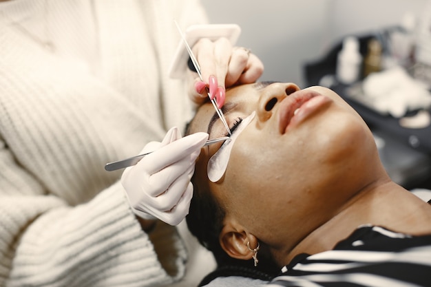 Master of eyelash extensions. African girl doing procedure. Woman on couch.