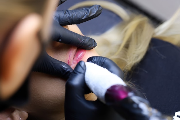 The master holds a tattoo machine and performs permanent makeup stretching the lips of the model