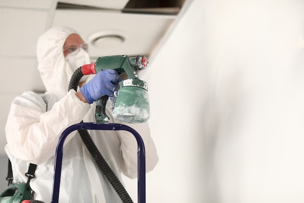 Master painter in protective suit is touching wall with spray gun