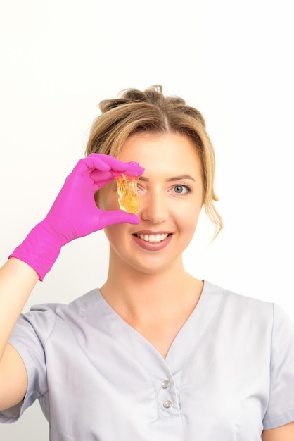 The master of sugar hair removal holds liquid yellow sugar paste covering her eye with wax for depilation on a white background