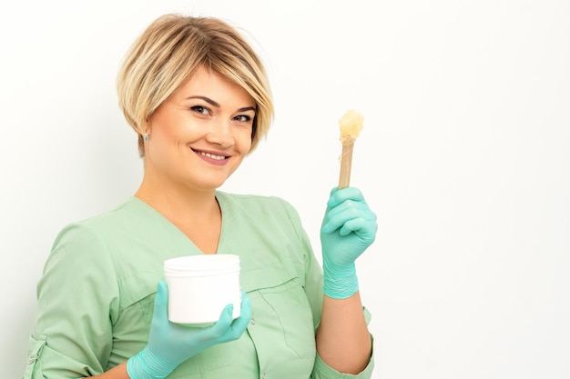 The master of sugar hair removal holds liquid yellow sugar\
paste wax for depilation with the jar on a wooden stick on a white\
background