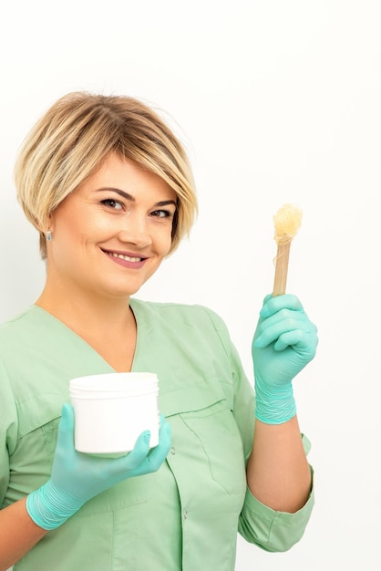 The master of sugar hair removal holds liquid yellow sugar paste wax for depilation with the jar on a wooden stick on a white background