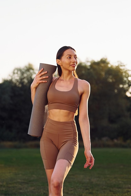 Mat is in the hands Young woman in yoga clothes is outdoors on the field