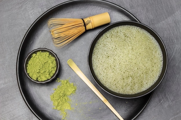 Matcha green tea in a bowl measuring spoon and matcha tea powder and a bamboo whisk on a black plate