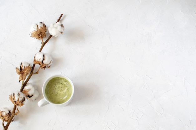 乾いた綿の枝の横にある白いカップの抹茶ラテ