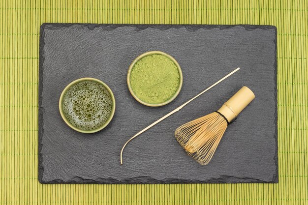 Matcha tea and Matcha tea powder in bowl bamboo whisk and tea spoon on stone board Green napkin made of wooden sticks Flat lay