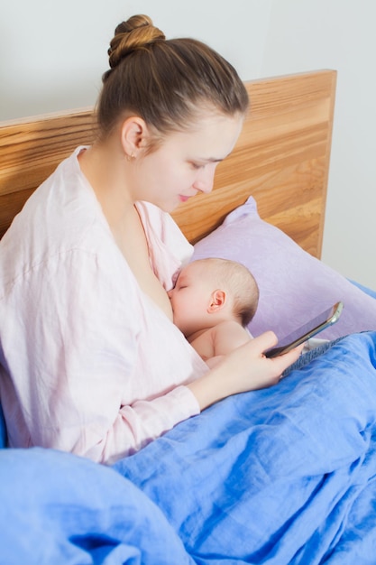 출산 휴가. 스마트폰으로 비디오를 보고 침실에서 어린 아기에게 모유 수유를 하는 어머니
