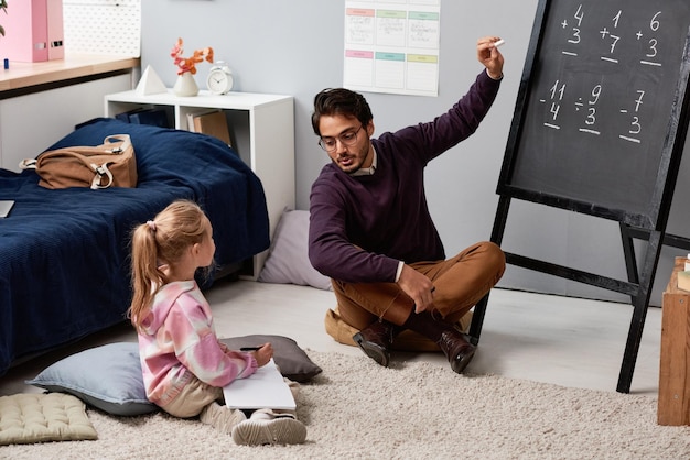 Math tutor sitting at blackboard and pointing at example while\
explaining rule to girl