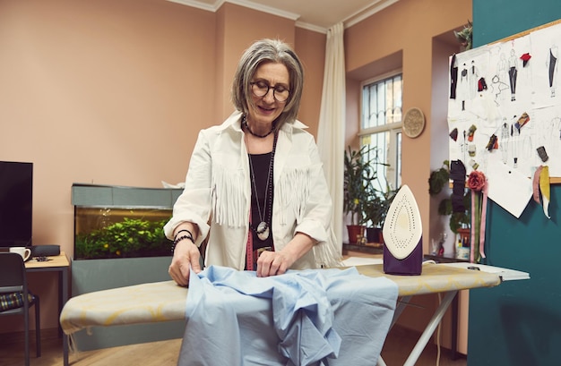 Mature 60 years old European woman fashion designer tailor ironing blue shirt from new collection in a tailoring atelier Seamstress sewer craftswoman needlewoman dressmaker fashion stylist