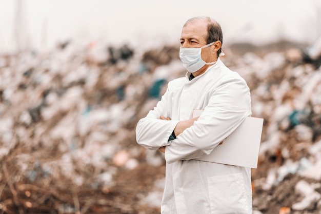 Mature adult Caucasian ecologist in white uniform and mask on face standing with crossed arms and holding clipboard under armpit.