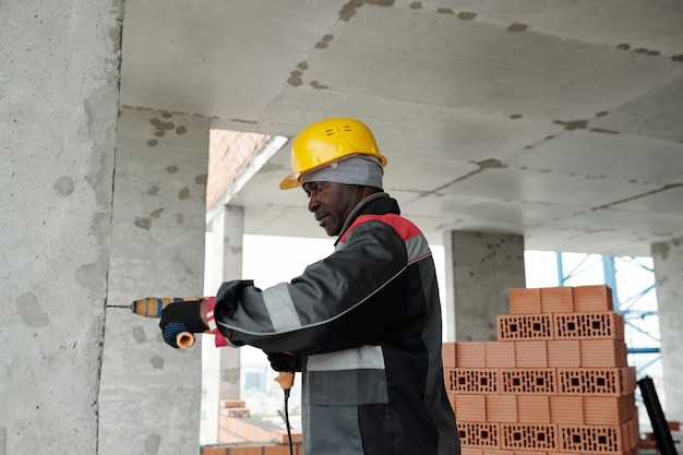 Mature african american builder in workwear using electric\
drill while working